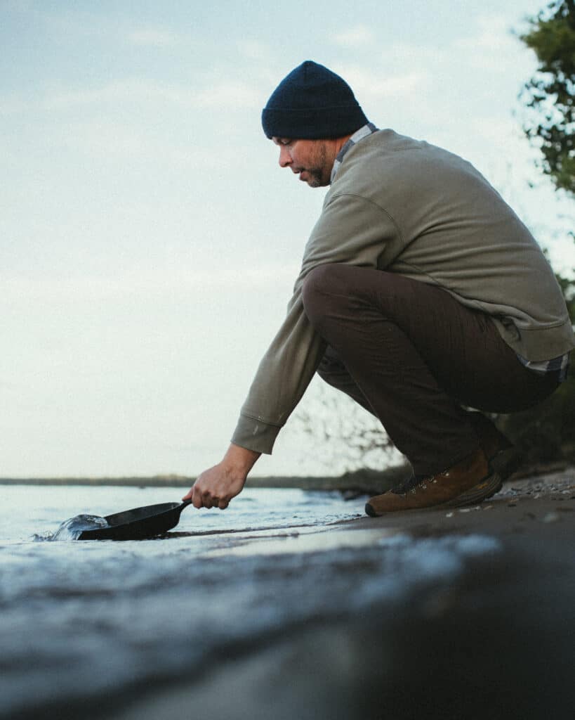 man cleaning cast iron skillet in lake
