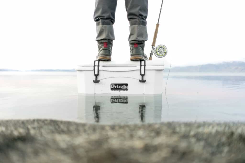 man standing on top of white grizzly cooler in the water with fly fishing rod and reel resting on the cooler lid