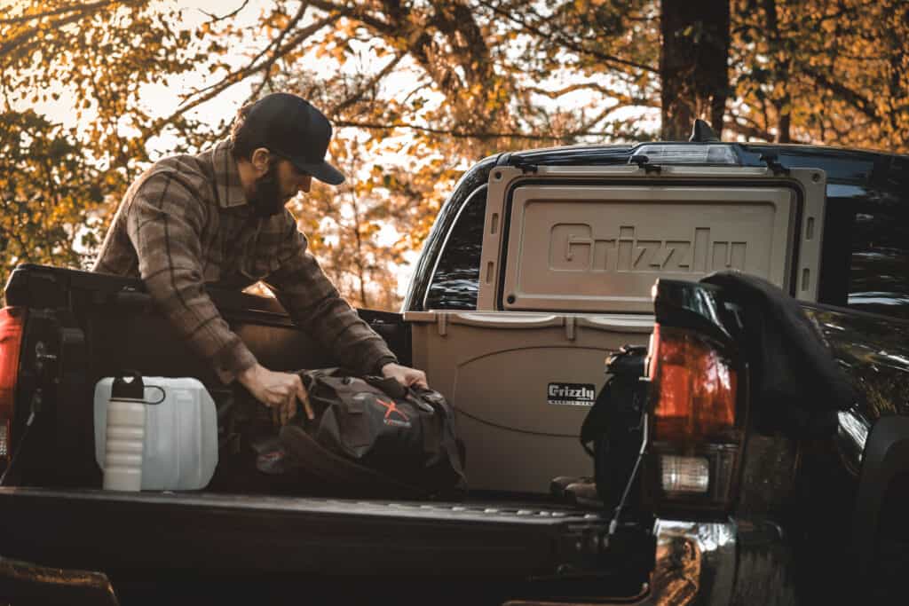 Grizzly 75 cooler with lid open sitting in back of pick up with man reaching into back of truck bed with hunting gear.